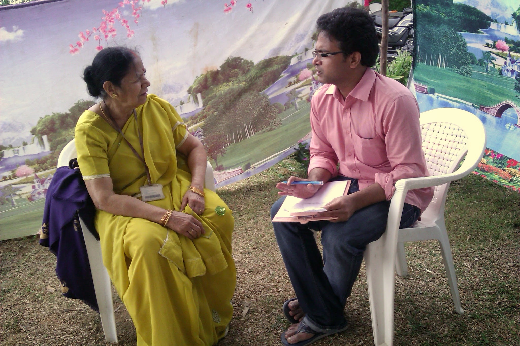 Dr. Suresh sitting on a plastic chair on the grass with a clipboard listening 1-on-1 to an elderly mother